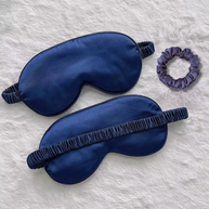 Two navy blue 22 momme mulberry silk sleep masks with elastic bands and a matching silk scrunchie on a white fuzzy surface.