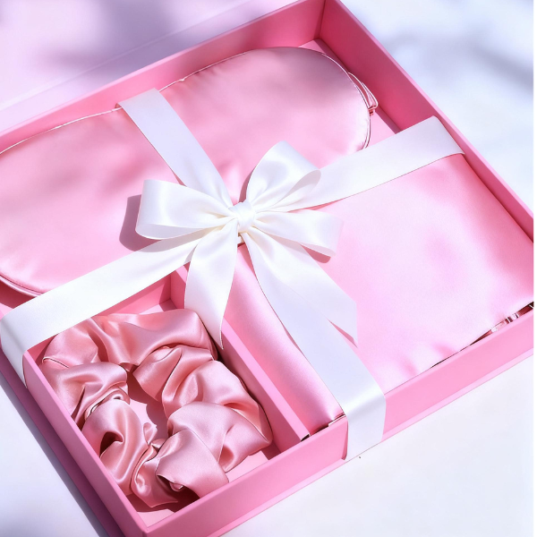 Pink satin pillowcase, eye mask, and two scrunchies in a pink gift box with a white ribbon.