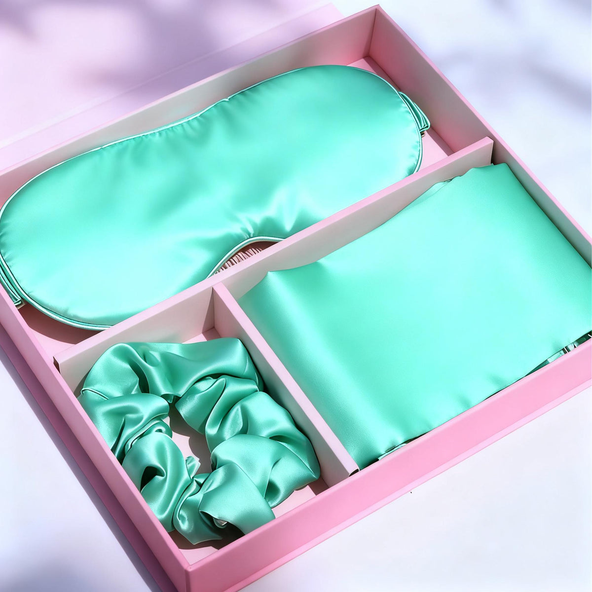 Mint green satin pillowcase, eye mask, and hair scrunchie in a pink gift box with ribbon.